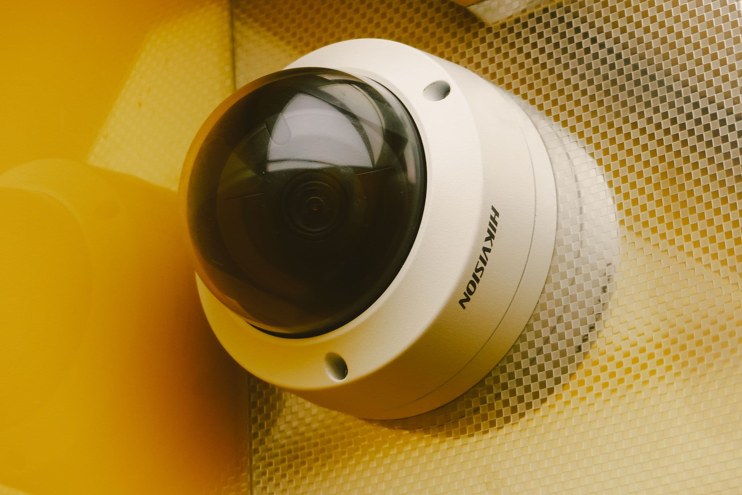 A dome security camera mounted against a yellow wall and metal mesh